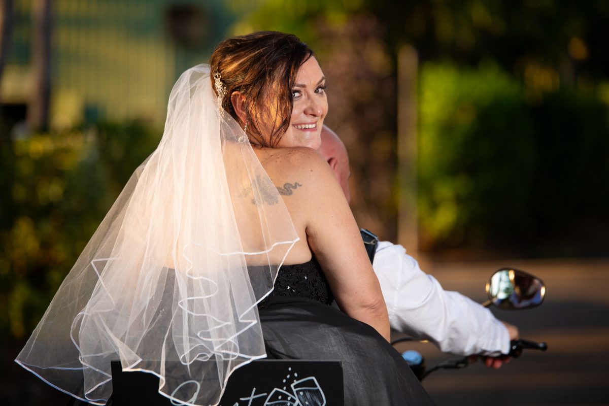 Bride looks back as wedding couple ride away on motorcycle.