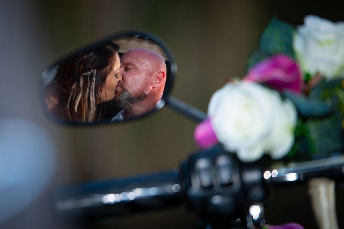 Reflection in motorcycle mirror of Bride and Groom kissing.