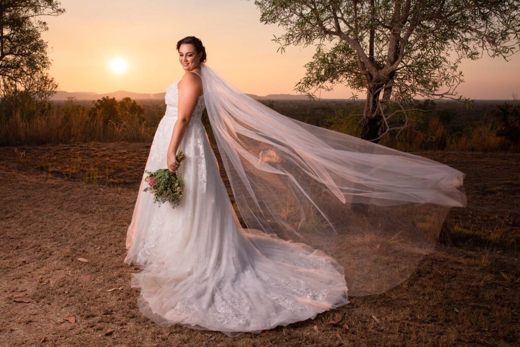 Bride poses with long veil and bouquet at sunset.