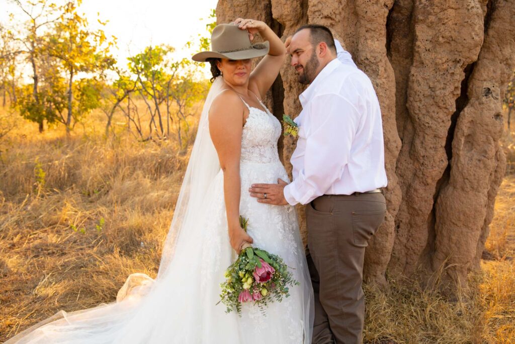 Bride wears groom&#039;s hat. they are leaning on a termite mound.